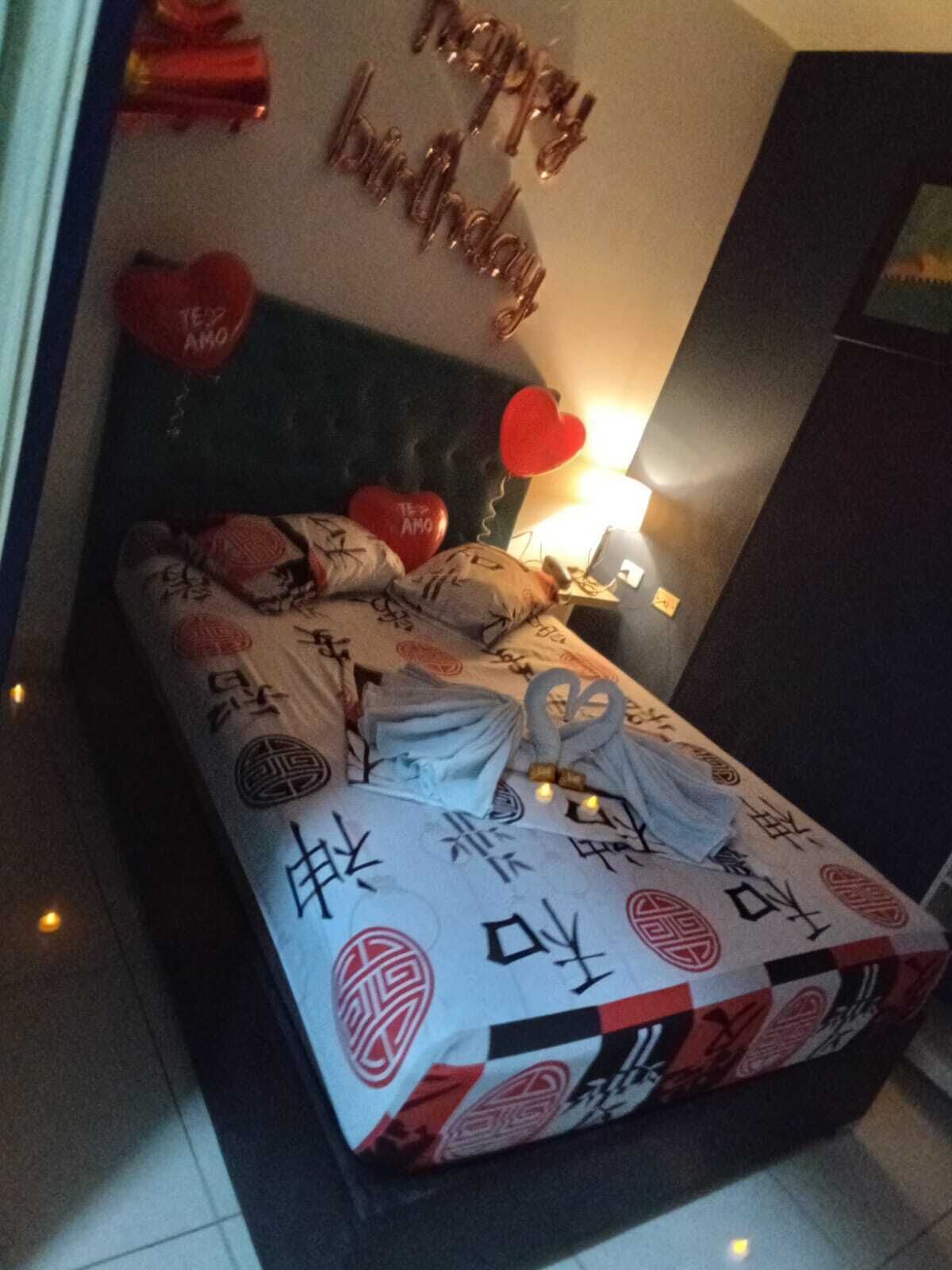 A neatly made bed with a patterned bedspread, a bedside lamp, and a wall decoration with 'Happy Birthday' balloons.