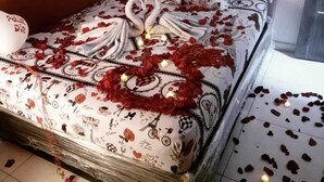 Una cama con colcha estampada de motivos florales y corazones, cabecera capitonada y almohada con forma de corazón y texto.