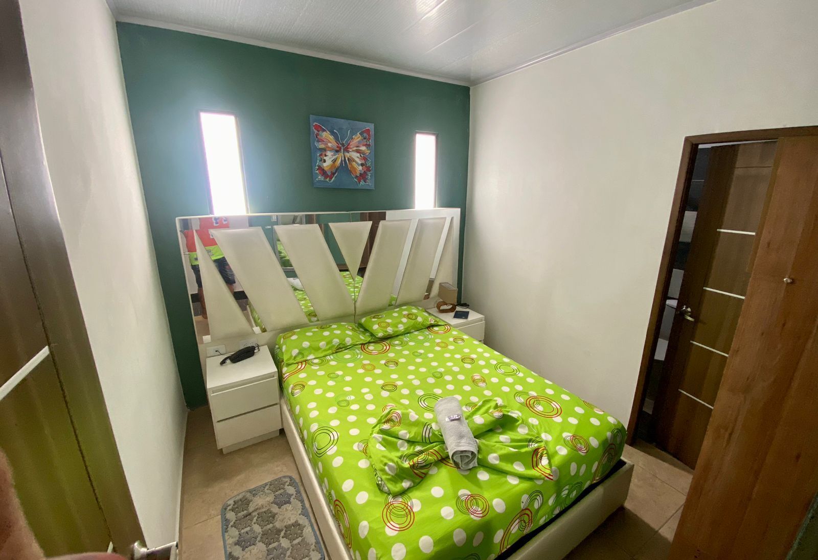 Ein Schlafzimmer mit Bett, Nachttisch, Wandregal, Tür und Fenster.