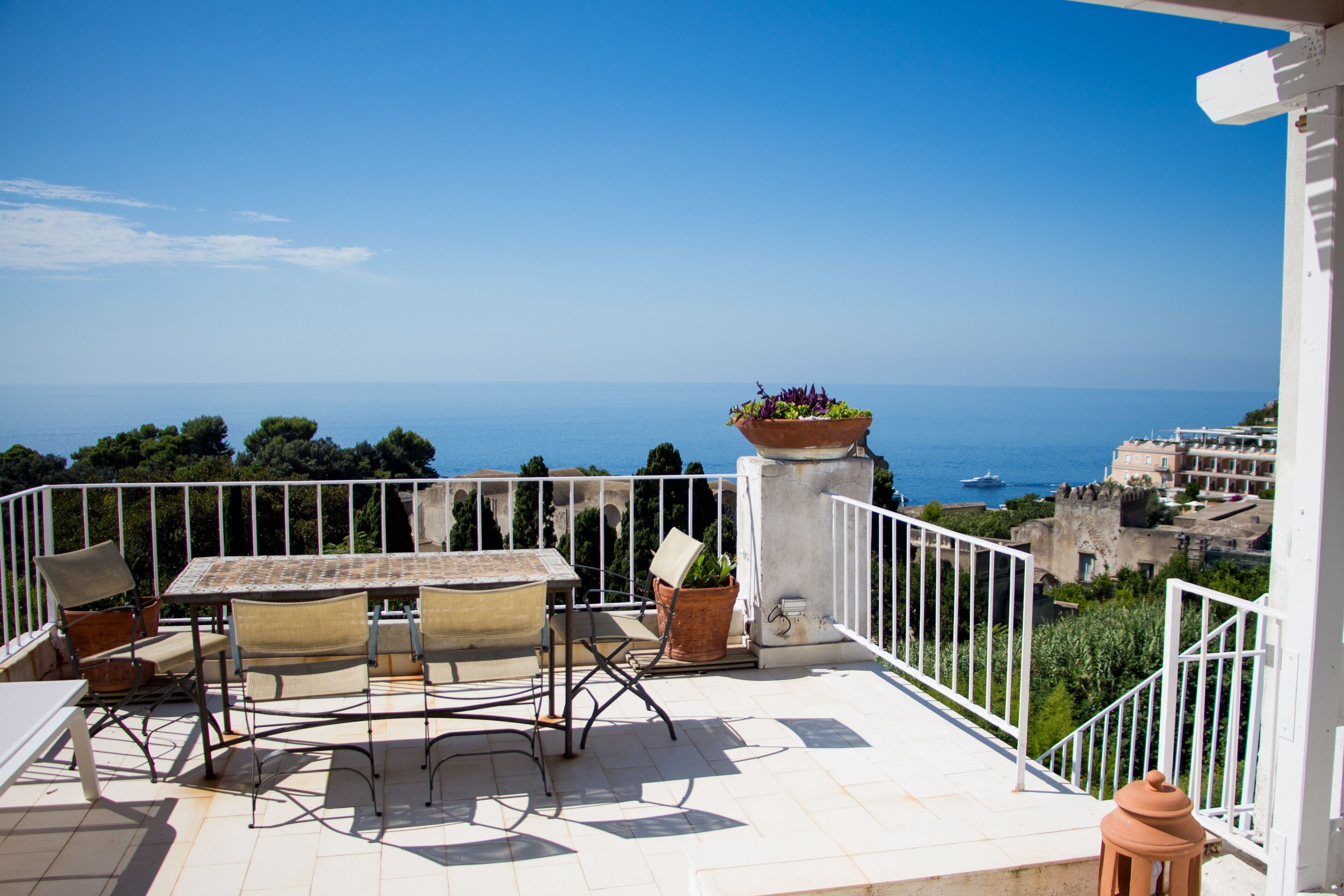 Une terrasse avec une table et des chaises, des plantes en pot et une vue sur la mer et les bâtiments.