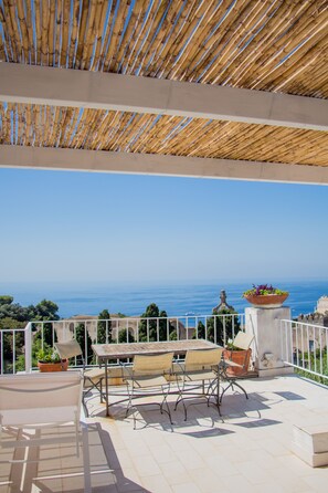 Um terraço com teto de bambu, mesa e cadeiras, e vista para o mar.