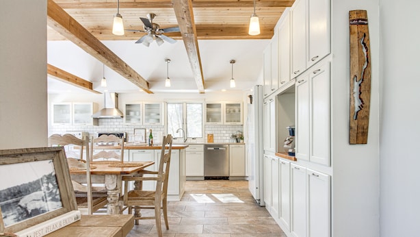 Una cocina con armarios blancos, techo de madera y comedor con mesa y sillas.