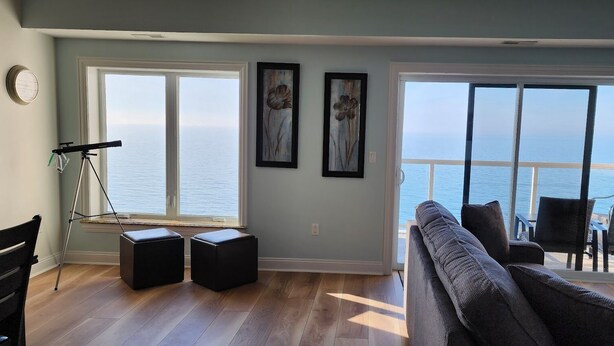 Ein Wohnzimmer mit großem Fenster zum Meer, einem grauen Sofa und zwei schwarzen Hocker.