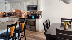 Eine Küche mit weißen Schränken, einer Granitabdeckung, einem Edelstahlkühlschrank, einer Mikrowelle, einem Kombi-Backofen und einem Essbereich mit Tisch und Stühlen.