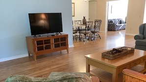 Una sala de estar con televisor sobre un mueble de madera, una mesa de comedor con sillas, un sofá y una silla de mimbre.