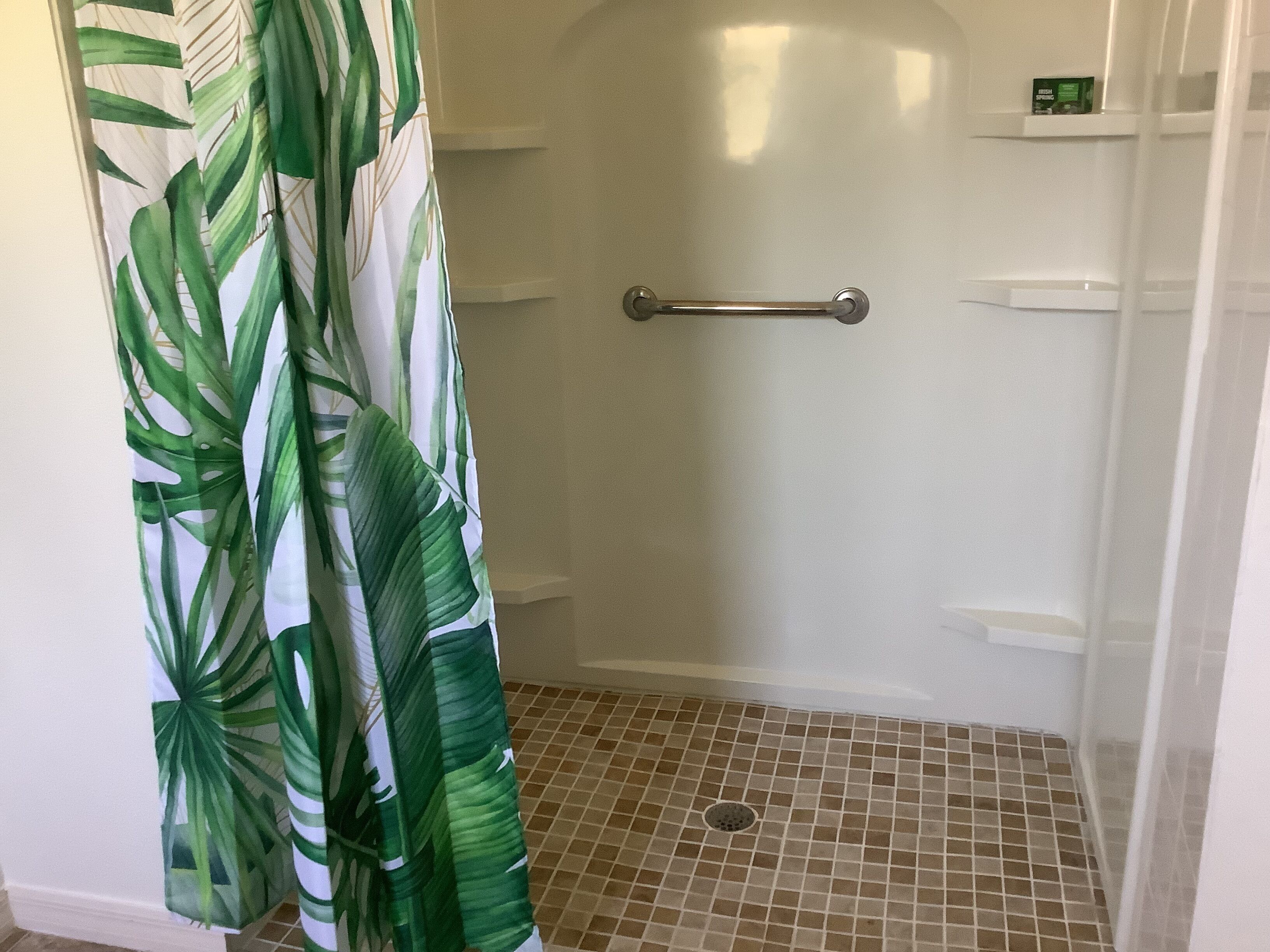 A shower area with a tiled floor, a handrail, and a shower curtain with a leaf pattern.
