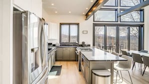 A modern kitchen with stainless steel appliances, a large island, and a view of the outdoors through large windows. - Lux Mountain Retreat w/ Heated Indoor Pool (Windham)