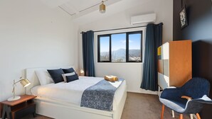 A bedroom with a bed, a chair, a nightstand, a lamp, a window with curtains, and an air conditioning unit.