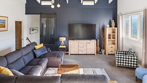 A modern living room with a dark blue accent wall, a large flat-screen TV, a grey sectional sofa, a wooden cabinet, and a patterned ottoman.