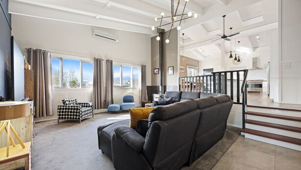A spacious living area with a large black sofa, a blue armchair, and a wooden coffee table. There are multiple windows providing natural light and a view of the outdoors.