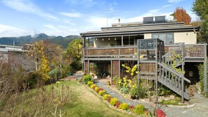 A modern two-story house with a balcony, a garden with various plants, and a view of mountains in the background.