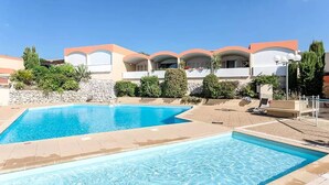 A swimming pool area with a clear blue pool, a stone wall, and a row of buildings with arched windows and a red roof. - Les Terrasses de Cassis (Cassis)