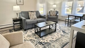 Una sala de estar con un sofá gris, un sillón a juego, una mesa de centro y un tapete estampado. Hay dos lámparas sobre mesitas auxiliares y un cuadro en la pared.