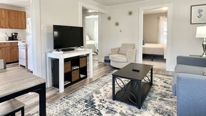 Una sala moderna con una televisión de pantalla plana, una mesa de centro, un sofá y una silla. Se ve un área de cocina a través de una puerta abierta.