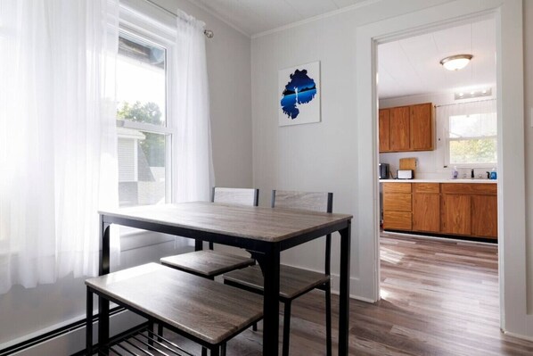 A dining area with a wooden table and benches, a view of the kitchen with wooden cabinets, and a painting on the wall.