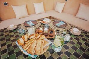 Disposição de mesa de pequeno-almoço com toalha quadriculada, bolos, bebidas e acompanhamentos.