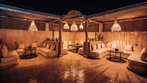 An outdoor lounge area with multiple sofas and tables, under a covered pergola with hanging lights. - Riad Villa Amood (Taouz)