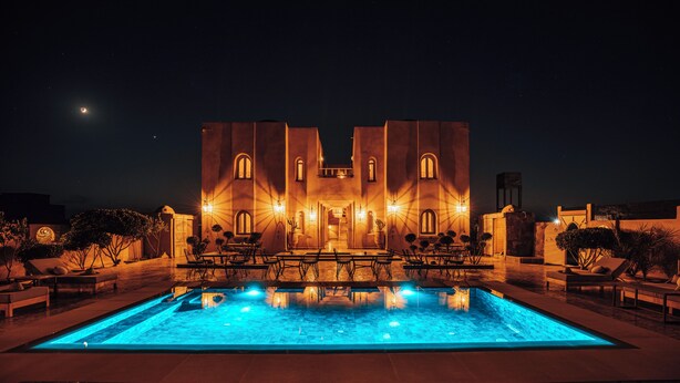 A pool area with a pool, lounge chairs, and a building with arched windows and lights. - Riad Villa Amood (Taouz)