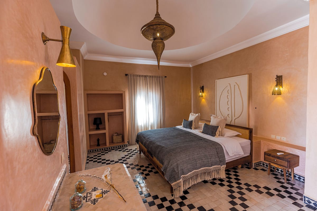 Une chambre avec un sol à carreaux, un lit recouvert d’une couverture grise, un luminaire suspendu et une lampe fixée au mur.