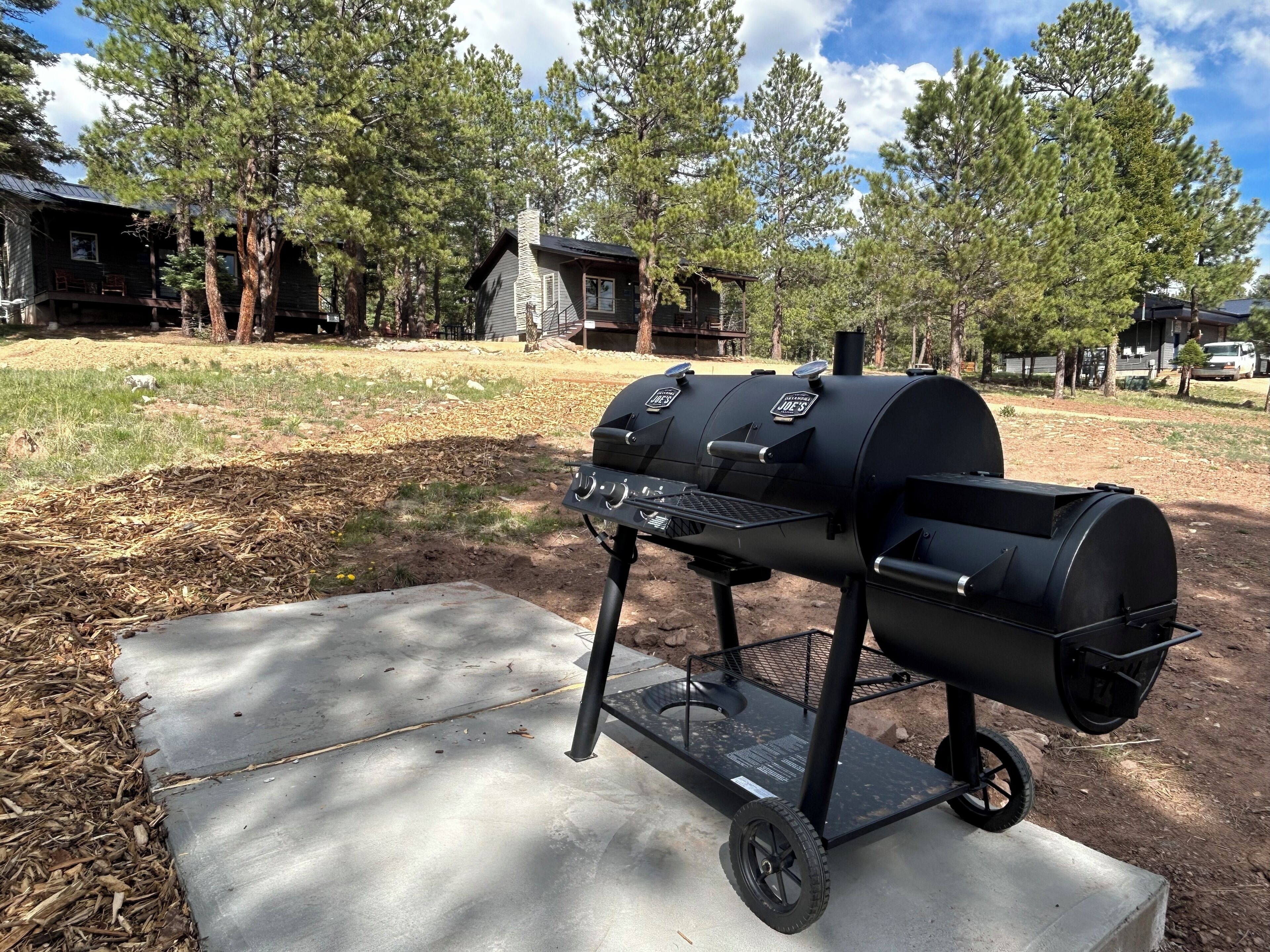 A portable barbecue grill with wheels, set on a concrete pad in a wooded area.