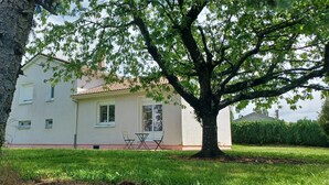 Il s’agit d’une maison à deux étages, avec une façade blanche et un toit recouvert de tuiles. Un grand arbre se trouve devant la maison, et un petit patio aménagé comprend une table et des chaises.