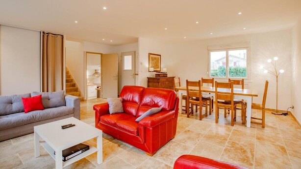 Un salon avec un canapé en cuir rouge, un canapé gris avec des coussins rouges, une table basse blanche et une table à manger en bois avec des chaises.