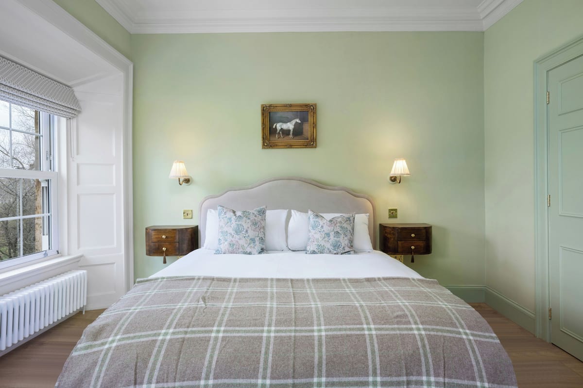 A bedroom with a large bed, two bedside tables, a window with a view, and a wall-mounted picture.