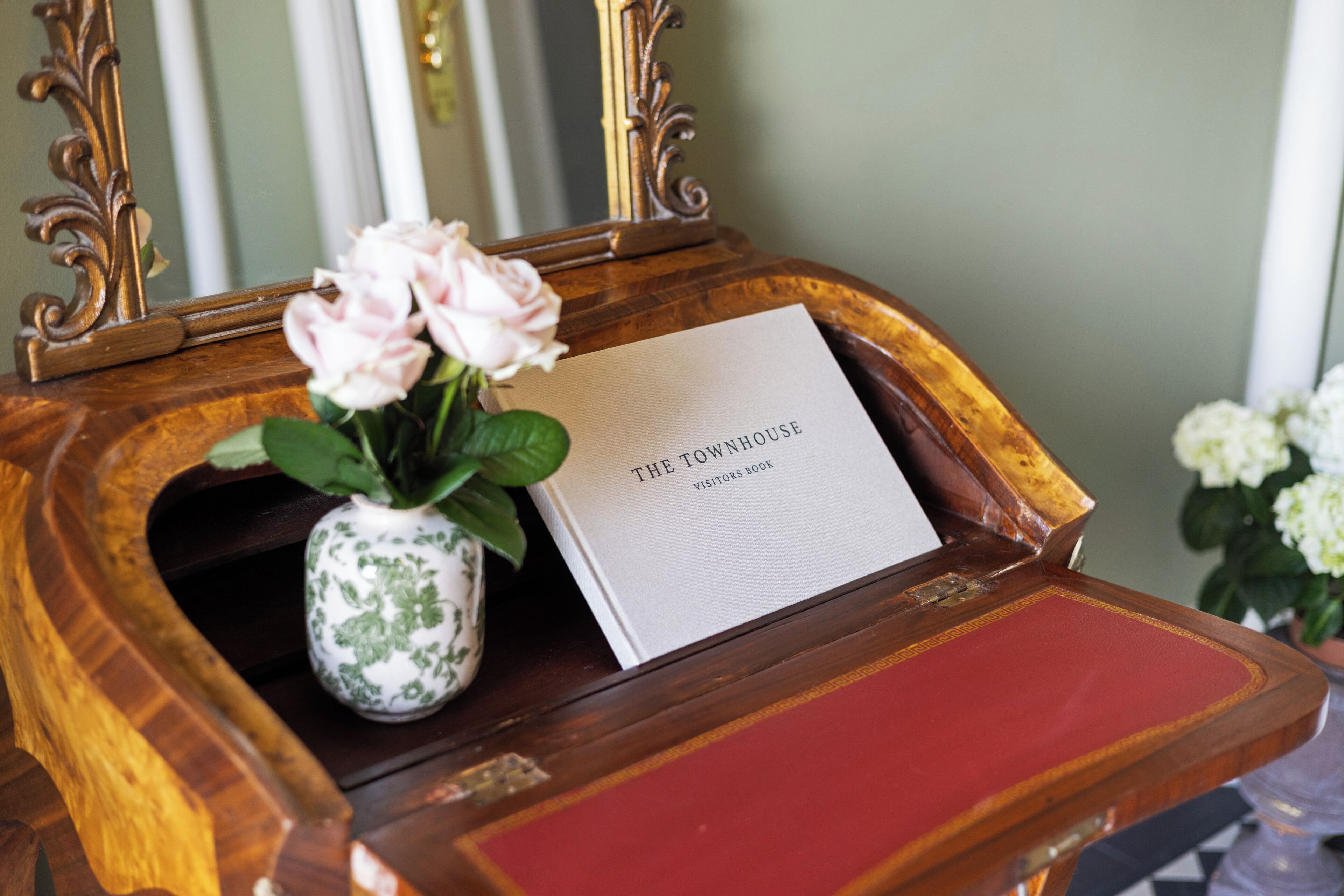 Una mesita de madera con un jarrón de rosas rosas y un libro blanco titulado 'The Townhouse Visitors Book'.