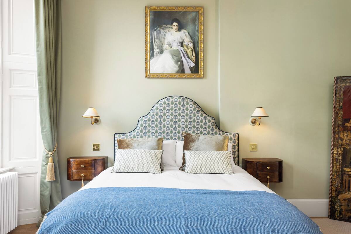 A neatly made bed with a blue bedspread, two matching pillows, and a headboard with a decorative pattern. There are two bedside tables with lamps and a framed painting above the bed.