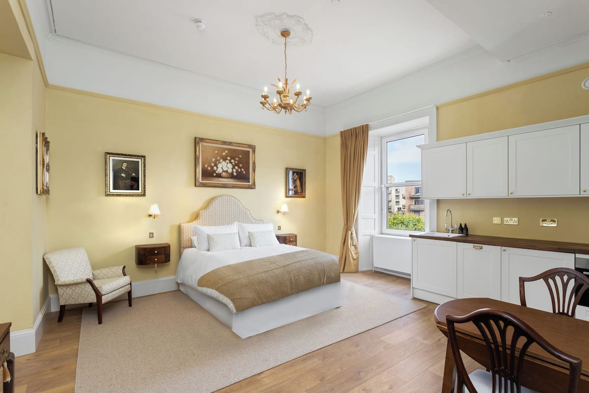 A bedroom with a large bed, bedside tables, a chair, a dining table, and a view of the city through a window.