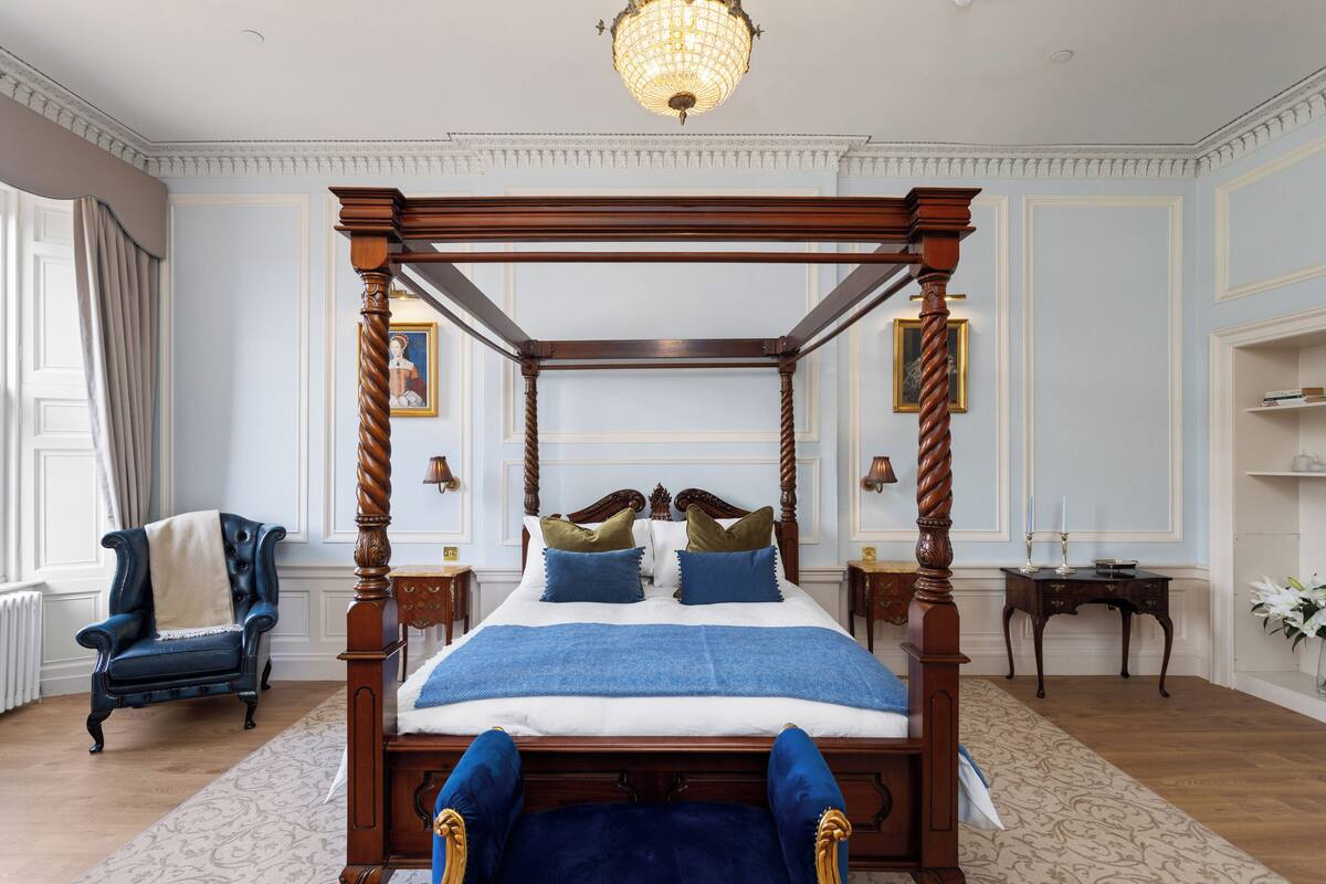 A four-poster bed with a wooden frame and a blue bedspread. There is a chair with a blue cushion and a white throw blanket. A chandelier hangs from the ceiling.