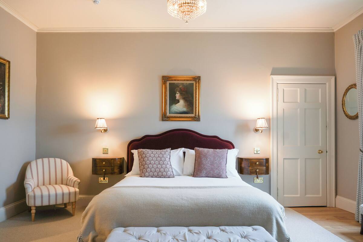 A bedroom with a large bed, two bedside tables, a chair, a door, and a chandelier.
