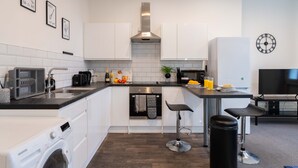 A modern kitchen with white cabinets, a stainless steel range hood, a built-in oven, a washing machine, and a bar area with stools. - 30% OFF Monthly | Family & Work Stays - Contact us for better rates! (Woodhouse)