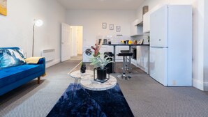A modern living room with a blue sofa, a coffee table with a plant, and a kitchen area with a white refrigerator. - 30% OFF Monthly | Family & Work Stays - Contact us for better rates! (Woodhouse)