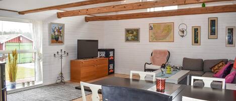 A cozy living room with a wooden ceiling, a television on a wooden cabinet, a dining table with chairs, and a sofa.