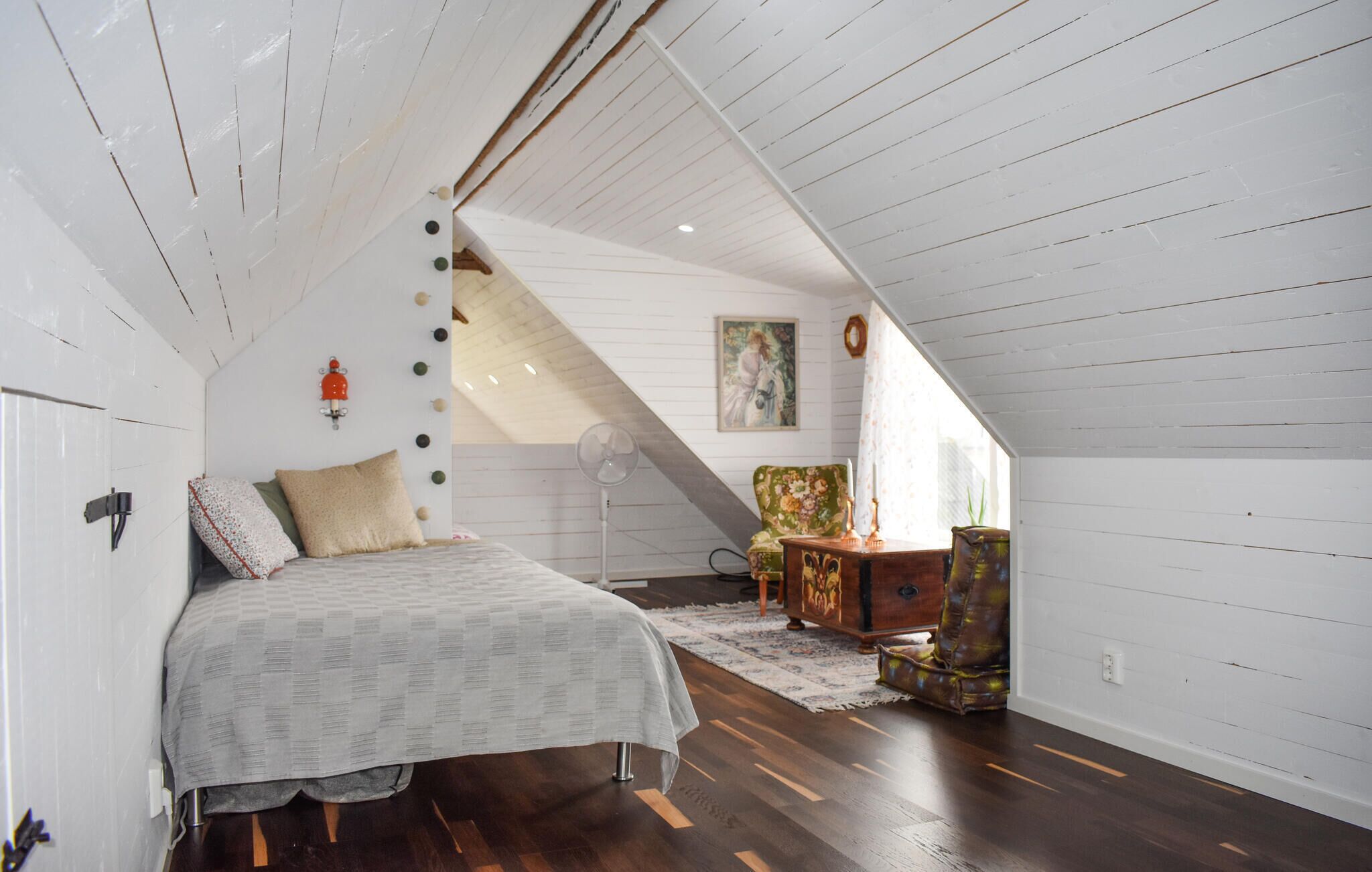 Une chambre avec un plafond en pente, un lit, une chaise, une commode et un tableau accroché au mur.