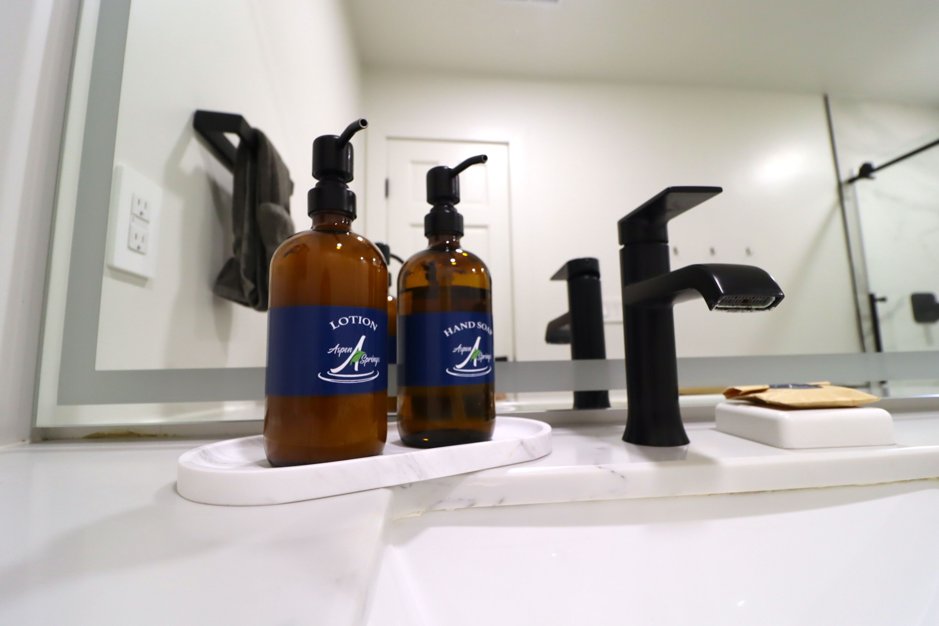 A bathroom with a white sink, two bottles of lotion, a black faucet, and a mirror reflecting the room.