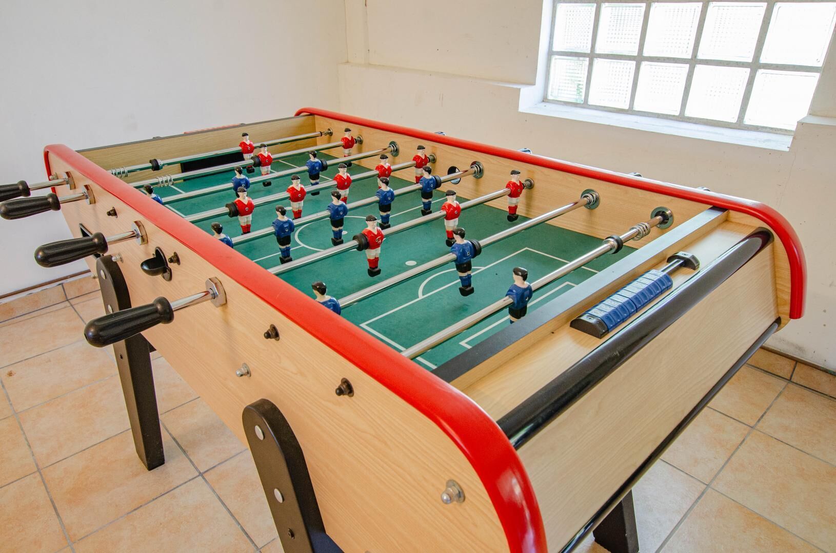 A foosball table with red and blue players in a room with tiled flooring and a window.