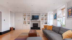 A modern living room with a grey sofa, a wooden coffee table, and a fireplace. There are white shelves with decorations and a television above the fireplace.