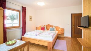A bedroom with a wooden bed, a television on a wooden shelf, a window with curtains, a radiator, and a dining table with a basket of apples. - Studio Apartment 'Peter Paul  Penegal' with Mountain View and Shared Terrace (Eppan an der Weinstrasse)