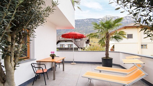 A balcony with a table, chairs, a red umbrella, and lounge chairs. - Studio Apartment 'Peter Paul  Penegal' with Mountain View and Shared Terrace (Eppan an der Weinstrasse)