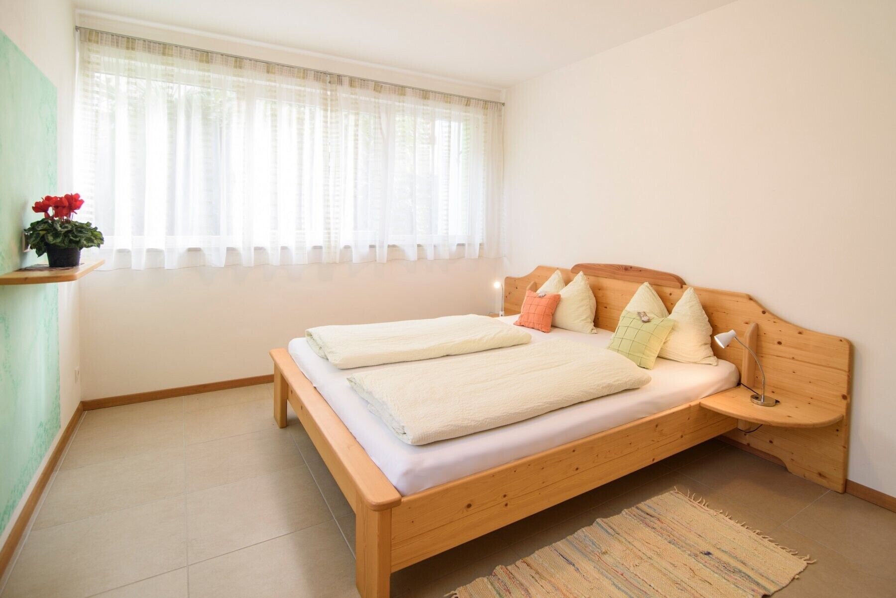 Habitación individual con una cama y cabecera de madera, una mesita de noche, una ventana con cortinas traslúcidas y una pequeña planta en maceta.