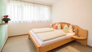 Habitación individual con una cama y cabecera de madera, una mesita de noche, una ventana con cortinas traslúcidas y una pequeña planta en maceta.