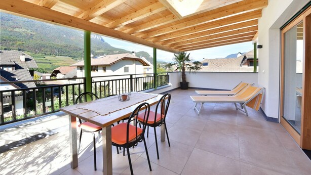 A covered balcony with a table and chairs, a lounge area with a sofa, and a view of the surrounding landscape.