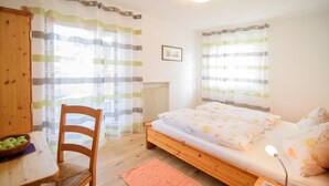 A single bed with a wooden frame, a small table with a basket of apples, a chair, and a radiator.