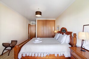 Une chambre d’hôtel avec un lit en bois, une table de chevet en bois, une chaise, une lampe et une porte.