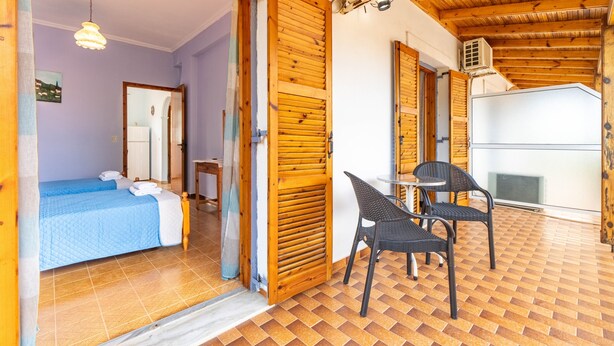 Una habitació amb un llit, una cadira, una taula i una porta oberta que dona a una altra habitació.