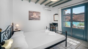 A bedroom with a bed, bedside lamps, a desk, a chair, and a view of the sea through a door.