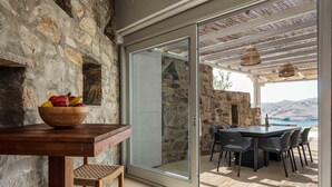A stone wall, a wooden table with a fruit bowl, a wicker chair, a glass door leading to an outdoor patio with a table and chairs, and a hanging wicker light fixture. - Villa 'Vagia' with Private Pool, Private Terrace and Air Conditioning (S???f??)