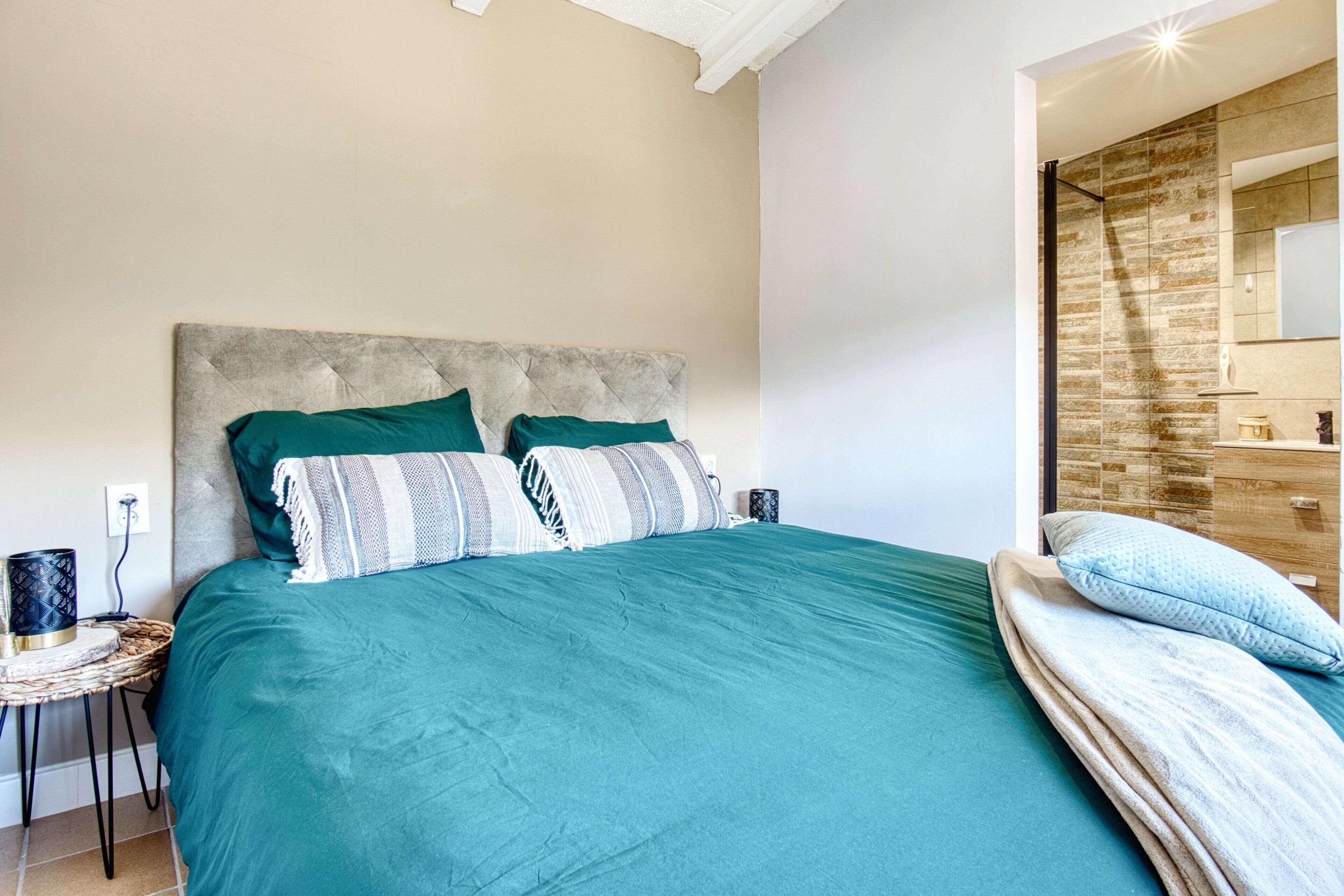 A bedroom with a large bed, a bedside table, a lamp, and a view of a bathroom with a tiled wall.